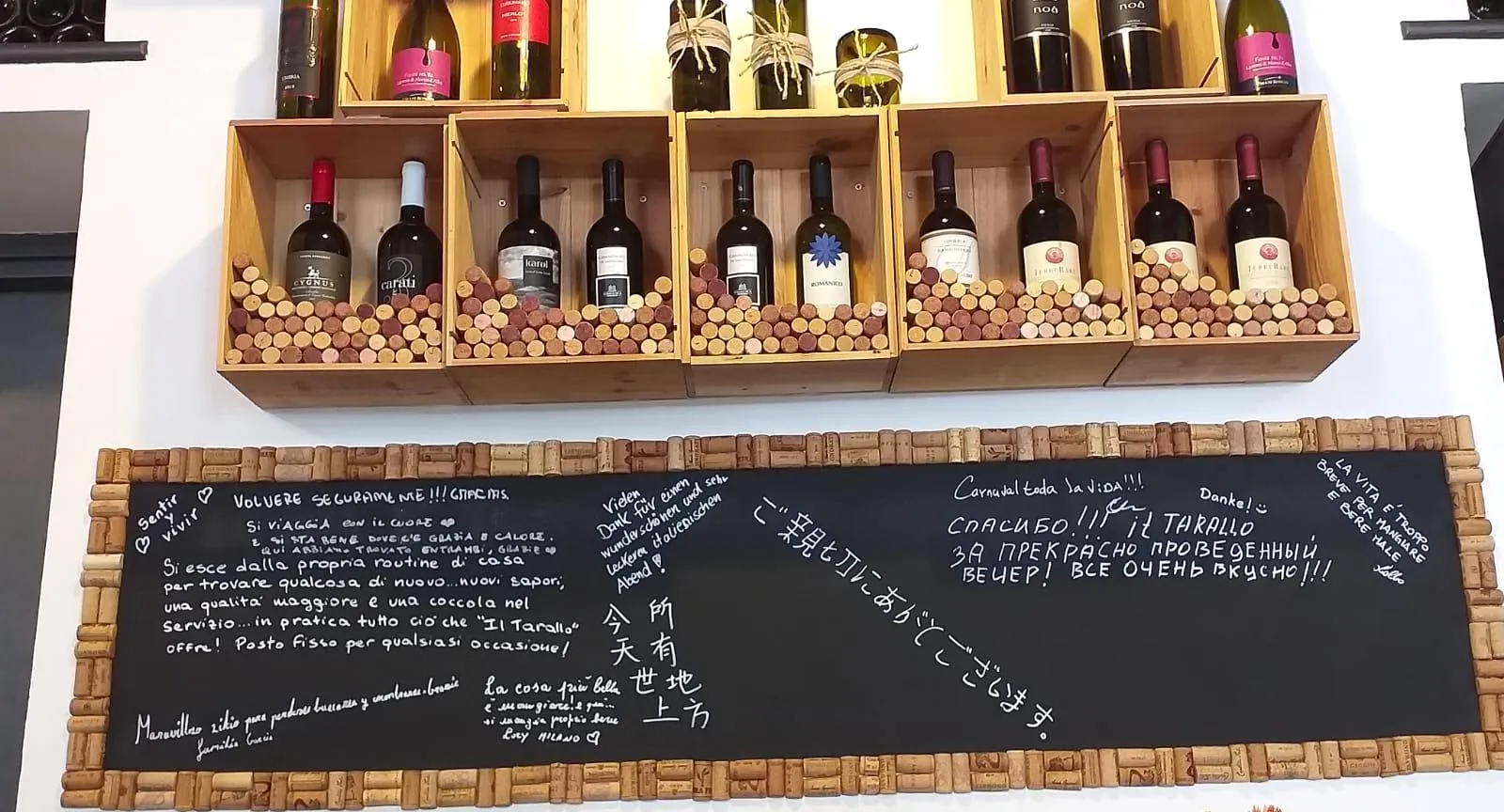 La lavagna con la cornice di tappi, piena di dediche dei clienti in tante lingue, sotto le cassette del vino